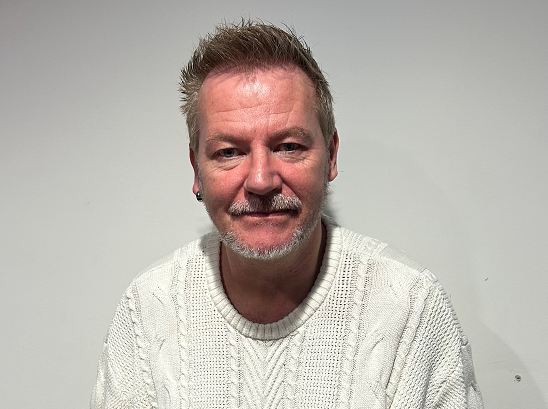 A person with short, light-coloured hair is wearing a white cable-knit sweater and dark pants. The person is standing against a plain, light gray wall with hands in pockets.