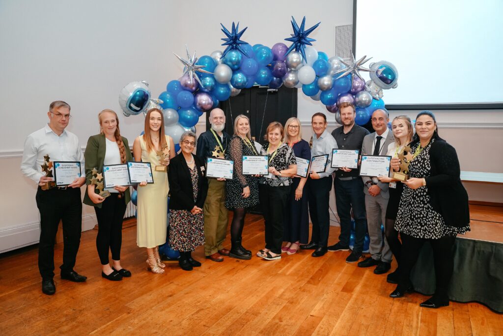 A group of people standing together celebrating their award. They are holding certificates and smiling at the camera in front of a blue and white balloon arch.